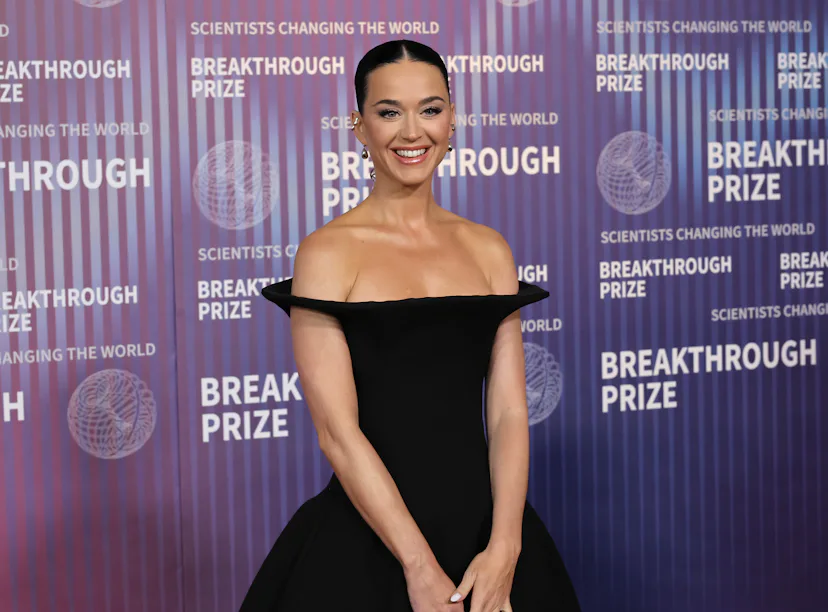 A woman in a black off-shoulder dress smiling at a Breakthrough Prize event, with a logo background.