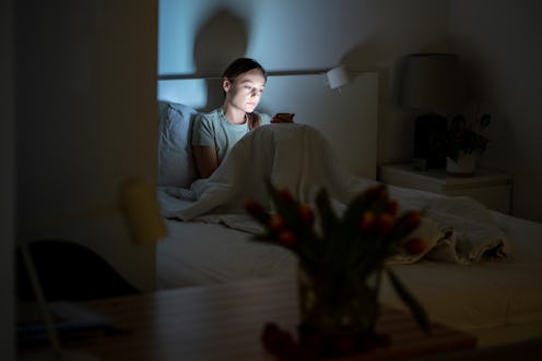 Sleepy tired woman sitting in bed, using smartphone late at night. Mobile phone illumination having ...