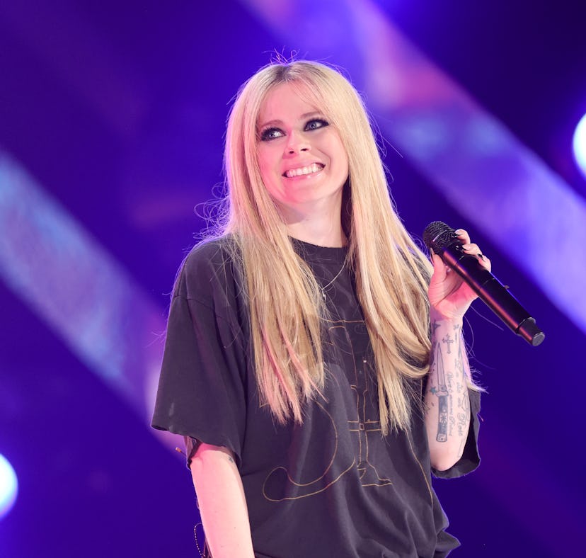 A female singer with long blonde hair performing on stage, holding a microphone, with bright lights ...