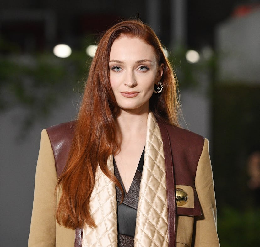 A woman with long red hair wearing a stylish layered outfit, including a quilted vest and brown leat...