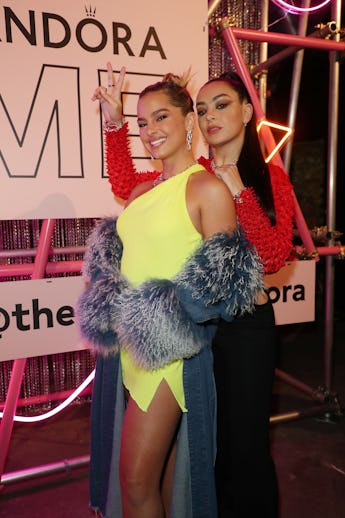 Two women pose at an event, one making a peace sign, both dressed in vibrant, fashionable outfits.