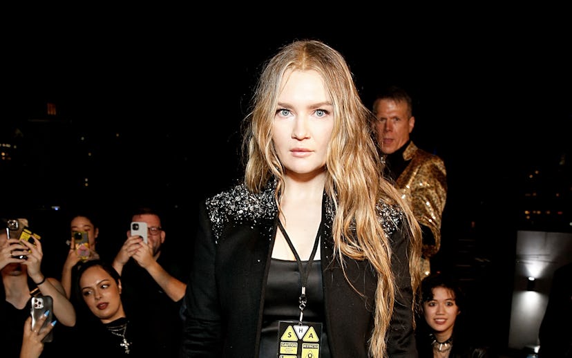 A woman in a black suit with tousled hair poses confidently at a crowded event, lit vividly against ...