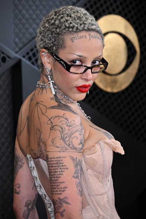 Woman with a short, curly hairstyle wearing glasses and a strapless dress, showcasing elaborate tatt...