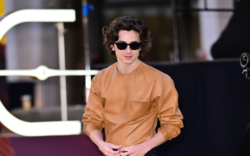 Person with curly hair wearing a camel color top, black leather pants, and sunglasses, walking down ...
