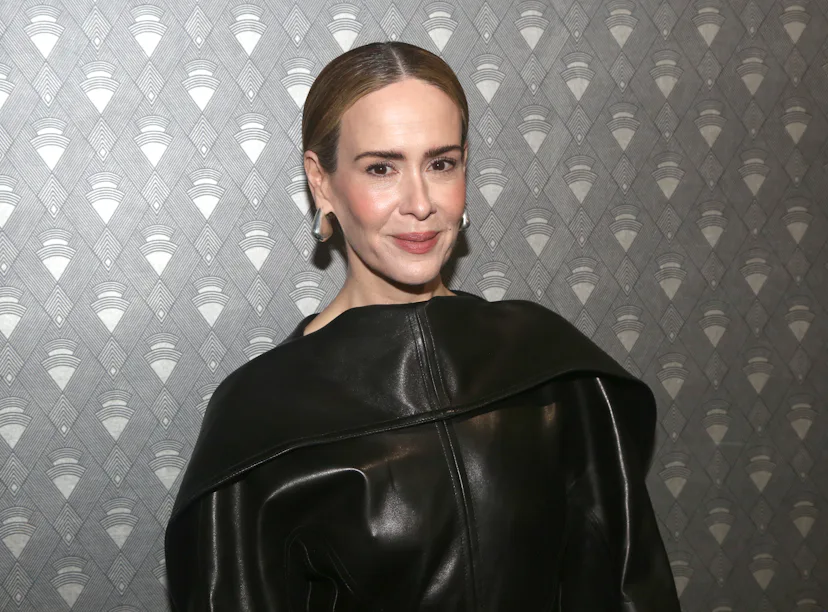 Woman with a confident smile wearing a stylish black leather jacket over a dress, standing against a...