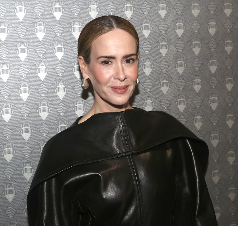 Woman with a confident smile wearing a stylish black leather jacket over a dress, standing against a...