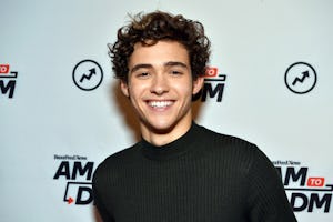 Young man with curly hair smiling in a black sweater at a red carpet event with logos in the backgro...