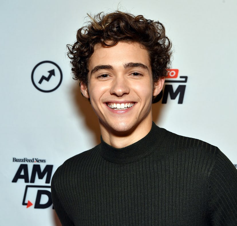 Young man with curly hair smiling in a black sweater at a red carpet event with logos in the backgro...