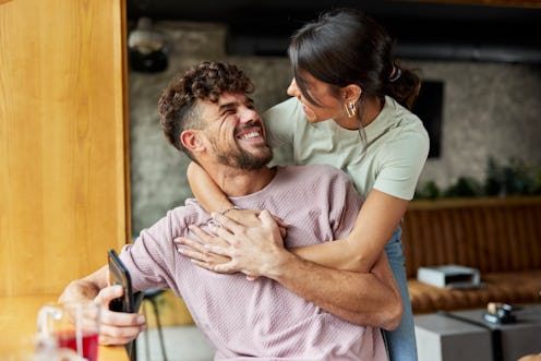 A joyful woman embraces a man in a wheelchair from behind, both smiling warmly in a cafe setting.