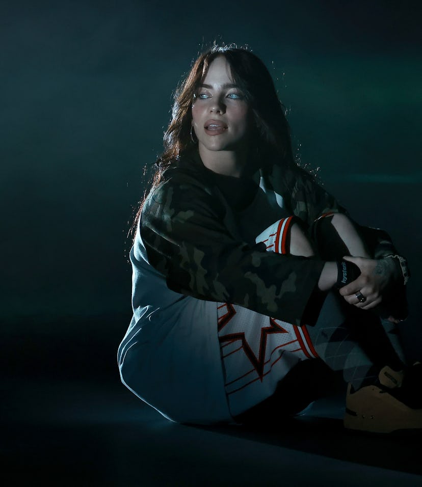 Woman wearing a camo jacket and graphic t-shirt sits on the ground under dim, moody lighting.
