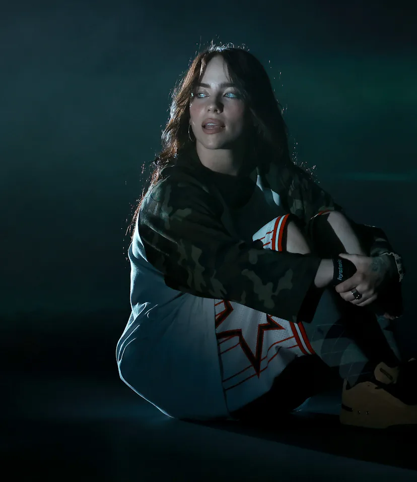 Woman wearing a camo jacket and graphic t-shirt sits on the ground under dim, moody lighting.