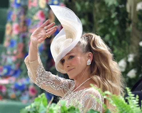 A woman in a bejeweled dress waves while wearing an elegant, large brimmed white hat at an ornate fl...