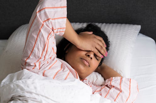 Woman lying in bed with her hand on her forehead, appearing unwell or distressed, wearing a striped ...