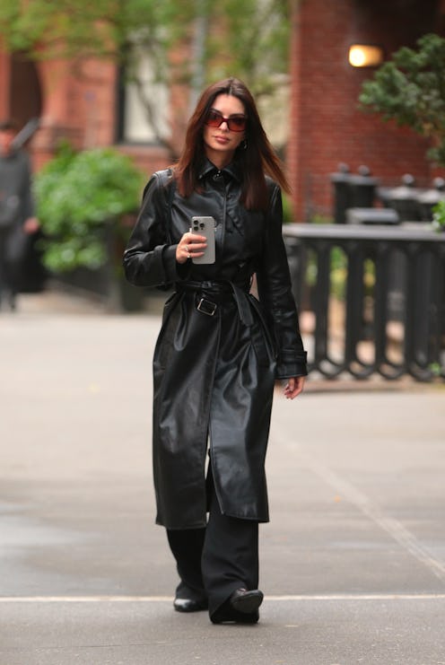 Woman in stylish black trench coat and sunglasses walks with smartphone in hand on a city sidewalk.