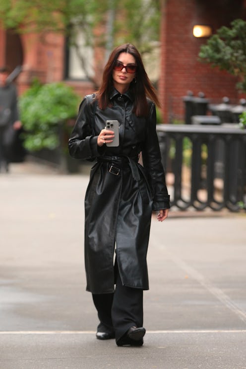 Woman in stylish black trench coat and sunglasses walks with smartphone in hand on a city sidewalk.