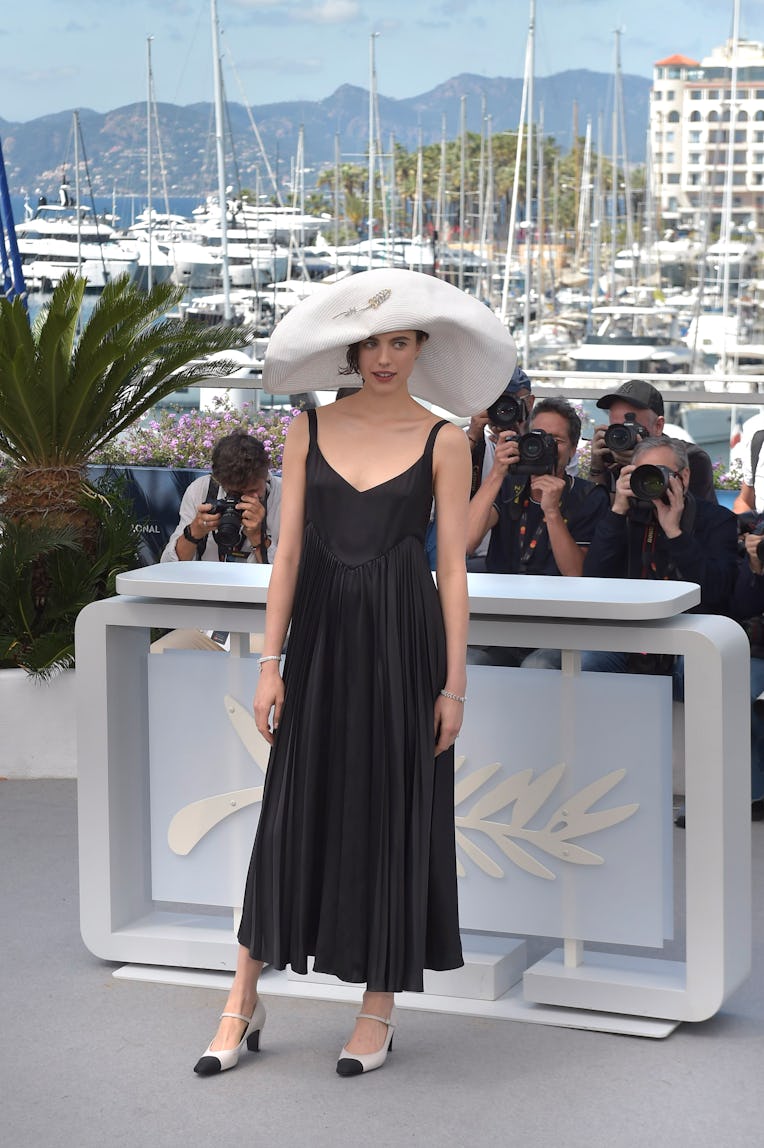Woman in black dress and large white hat poses in front of photographers at a sunny harbor event.