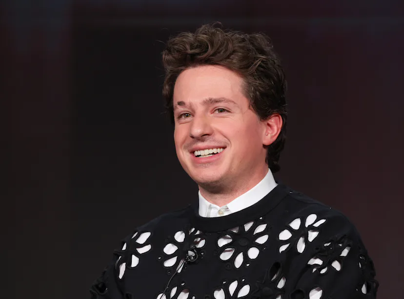 A man with curly hair smiling in a black shirt with white floral patterns, sitting against a blurred...