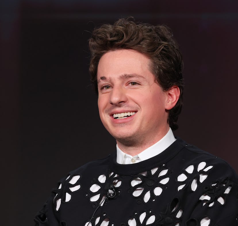 A man with curly hair smiling in a black shirt with white floral patterns, sitting against a blurred...