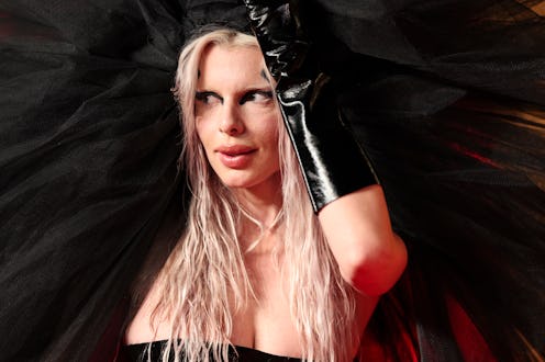 A woman with blonde hair, dressed in a black and red outfit, gazes intently while adjusting a long g...