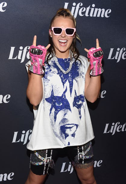 A person smiling broadly, posing with peace signs, wearing pink sunglasses and fingerless gloves, in...