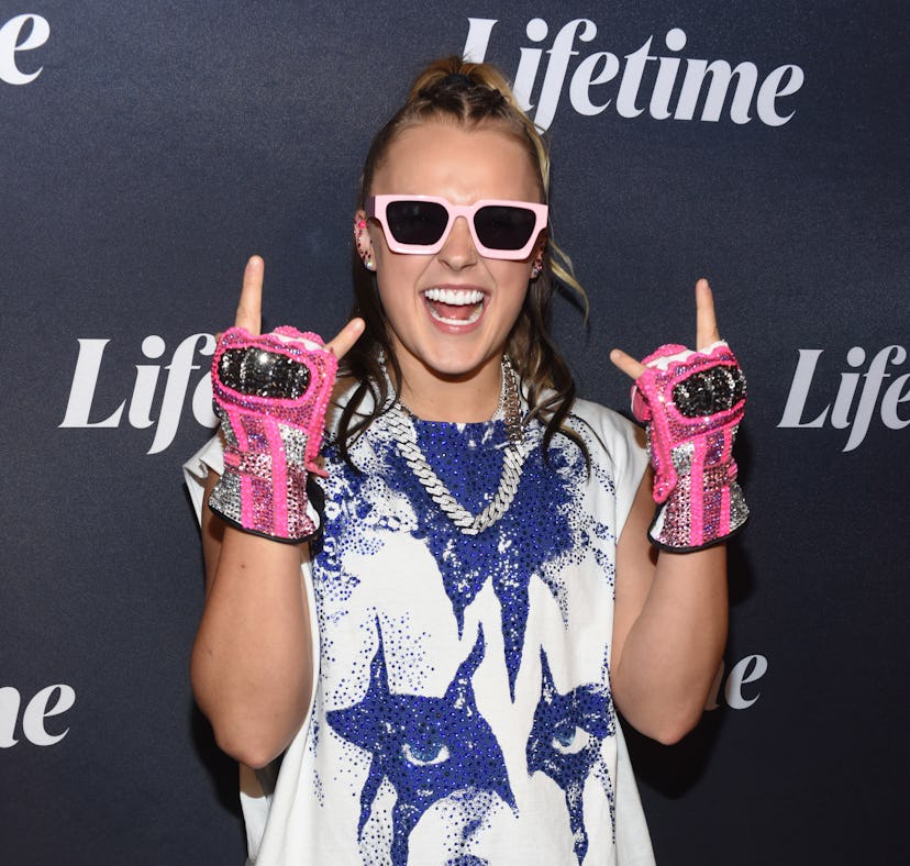 A person smiling broadly, posing with peace signs, wearing pink sunglasses and fingerless gloves, in...