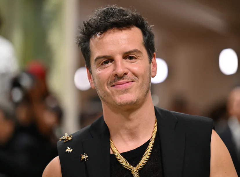 Man with a confident smile wearing a black sleeveless top adorned with star pins and multiple neckla...