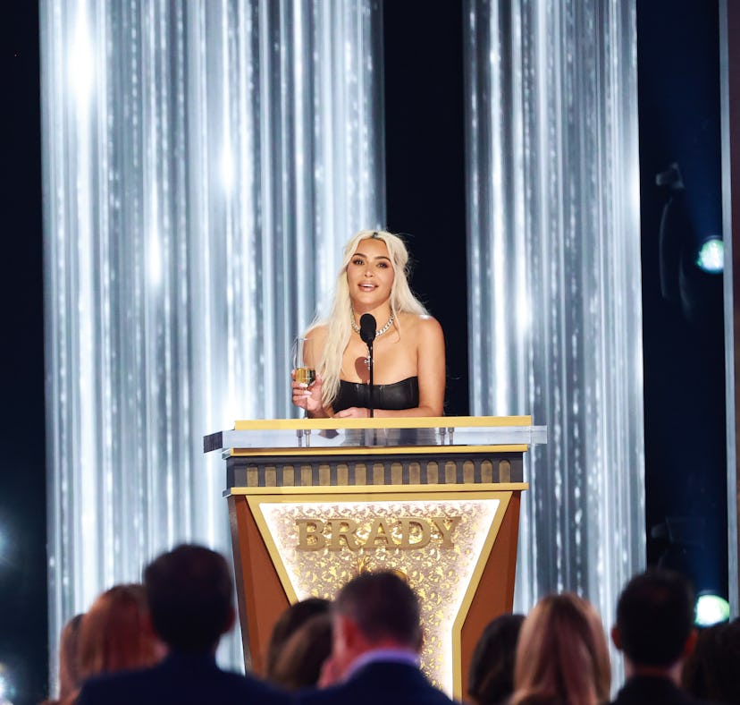 A woman speaks at a podium on a stage adorned with vertical light strands, addressing an audience at...