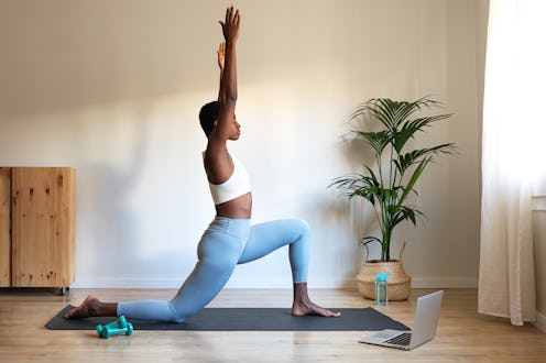 Woman practicing yoga in a bright room, using a mat and laptop for guidance, with plants in the back...