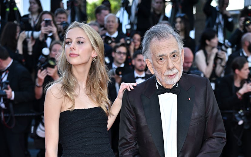 Young woman in a strapless black gown and elderly man in a tuxedo with a cane on the red carpet.