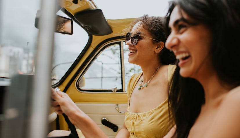 Two teenage girls driving a yellow car and laughing at corny jokes