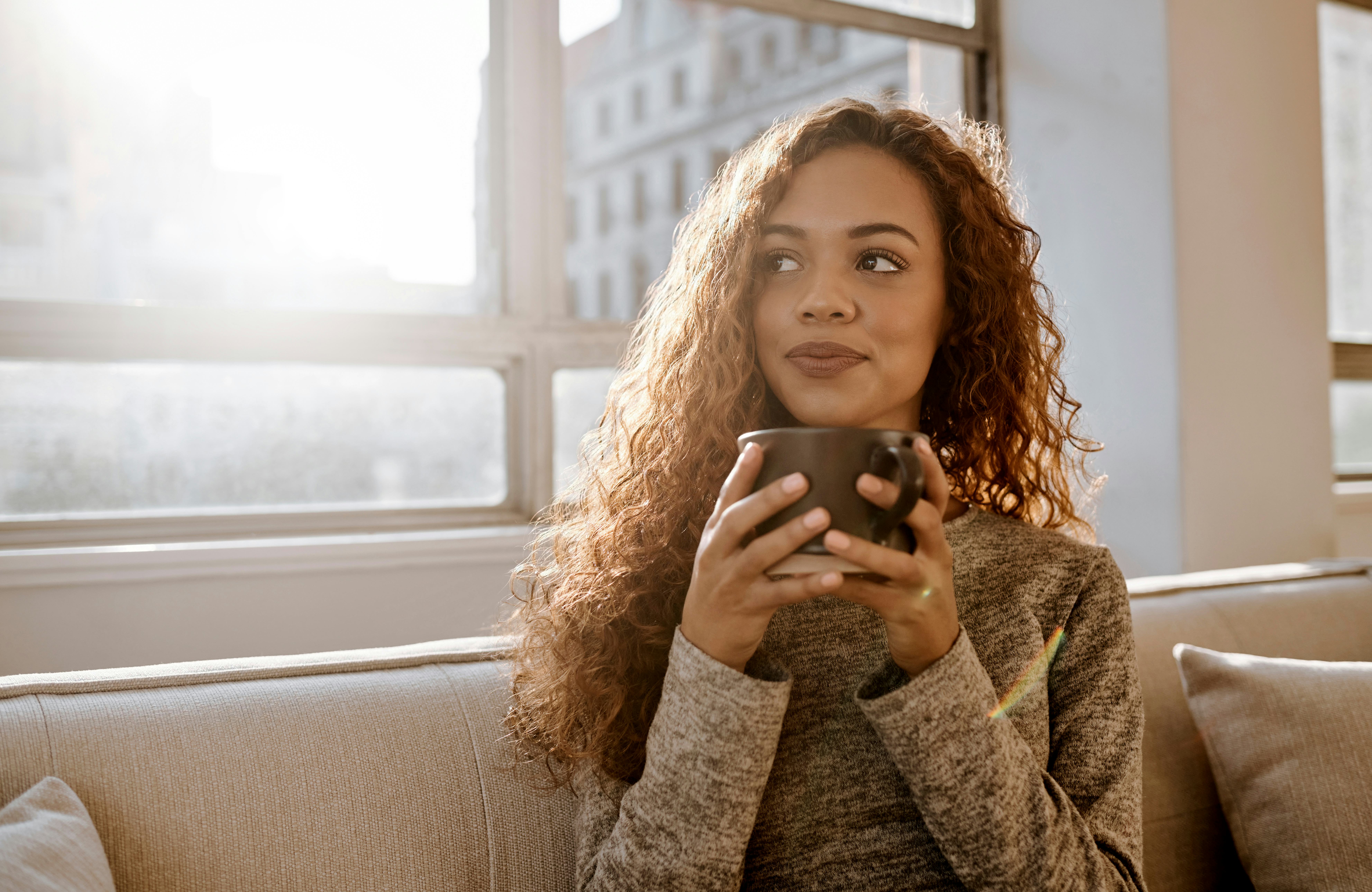 A young woman is sitting on a sofa thinking up some good jokes about coffee as she drinks her coffee&hellip;
