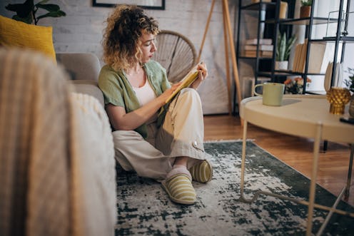 Casual young woman reads intently, sitting on the floor in a well-lit, stylish living room decorated...