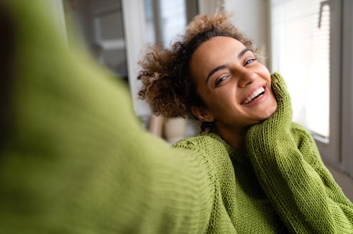 Beautiful smiling mixed-race woman is taking a selfie at home wearing an oversized green sweater.