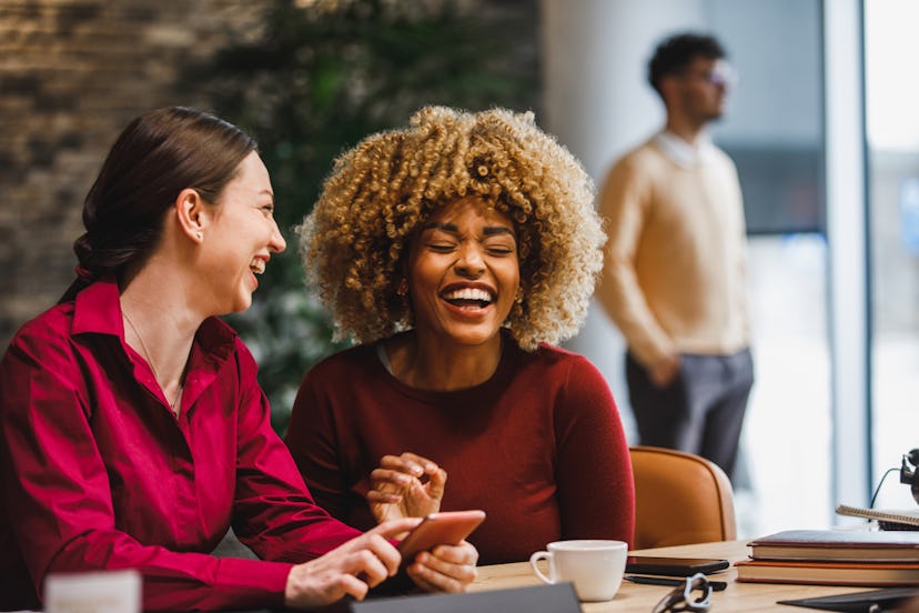 Two friends sitting in the office and laughing together while looking at good jokes on a smart phone…