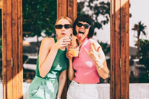 Two friends wearing fancy jewelry and sunglasses are having fun outside the cafe. Besties are drinki...