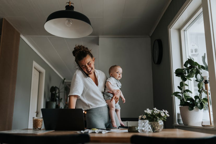 A young woman working from home and taking care of her baby, laughing at good jokes about working fr…