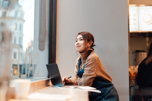 Young Asian woman looking through store window while working with laptop in cafe. Small business and...
