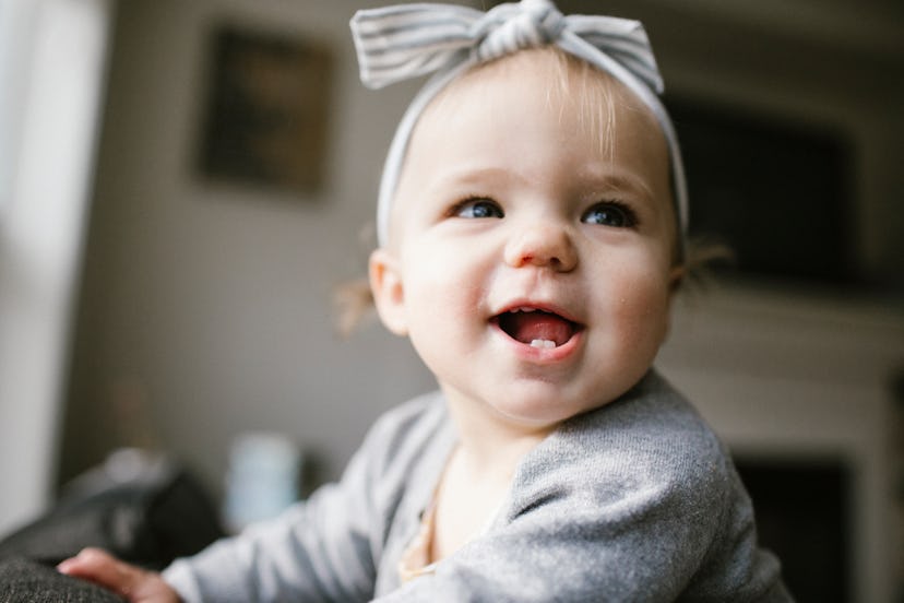 A baby with a trendy girl name in a bow smiling with two bottom teeth.