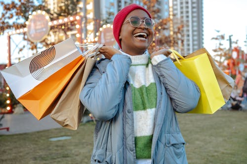 Joyful woman in a beanie and glasses carrying multiple shopping bags at an outdoor festive market.