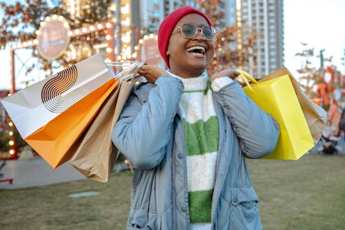 Joyful woman in a beanie and glasses carrying multiple shopping bags at an outdoor festive market.