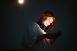A woman with red hair and headphones looks at her smartphone, illuminated by the phone's light in a ...