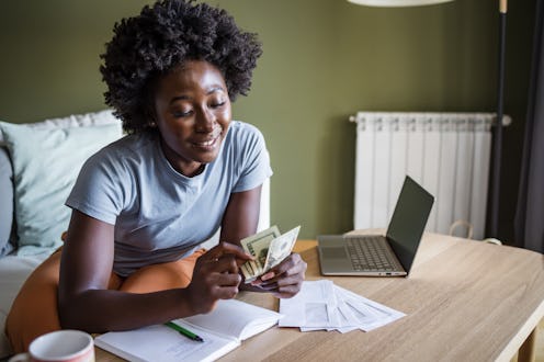 Woman counting money at a desk with a laptop and documents, smiling in a green-walled room.