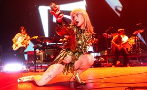 Female singer in a gold outfit performs on stage with a microphone, kneeling, with band members in t...