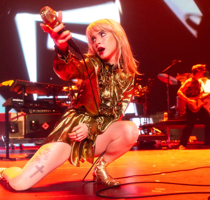 Female singer in a gold outfit performs on stage with a microphone, kneeling, with band members in t...