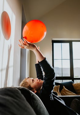 A little boy plays with a red balloon in a sunny room. He throws it up in the air and catches it aga...