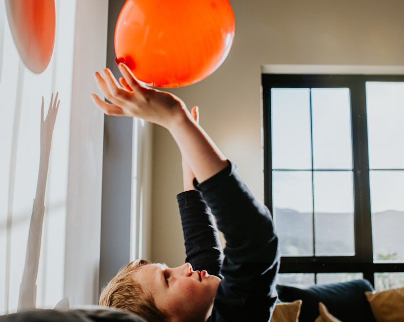 A little boy plays with a red balloon in a sunny room. He throws it up in the air and catches it aga...