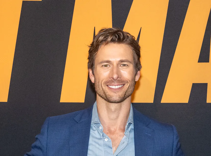 Man in blue suit and open-neck shirt smiling at a movie premiere against an orange and black backdro...