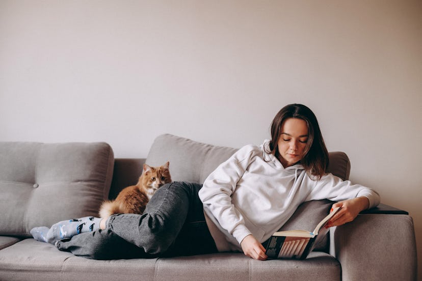 A woman reading a book of unique cat names on the couch with her cat lying on her legs.
