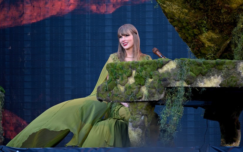 Singer in green dress performs on a moss-covered piano on an outdoor stage, with a digital screen ba...