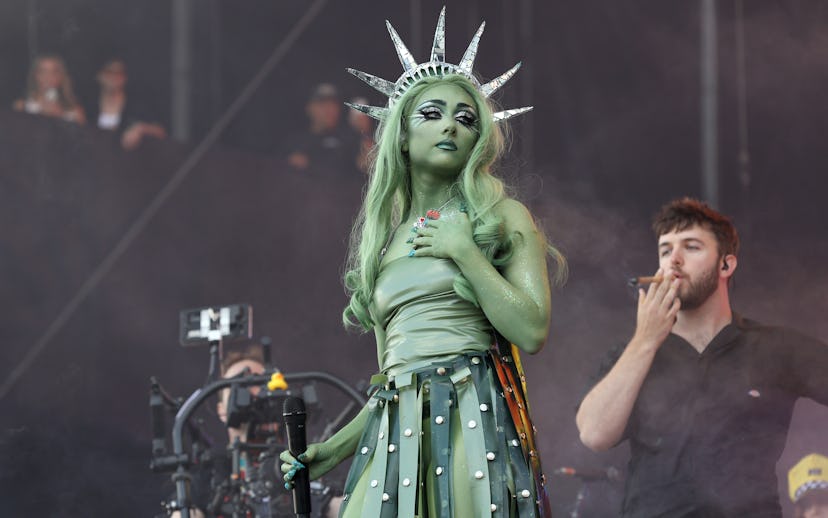 Performer dressed as the Statue of Liberty, with green skin and crown, on stage at a music event, wi...